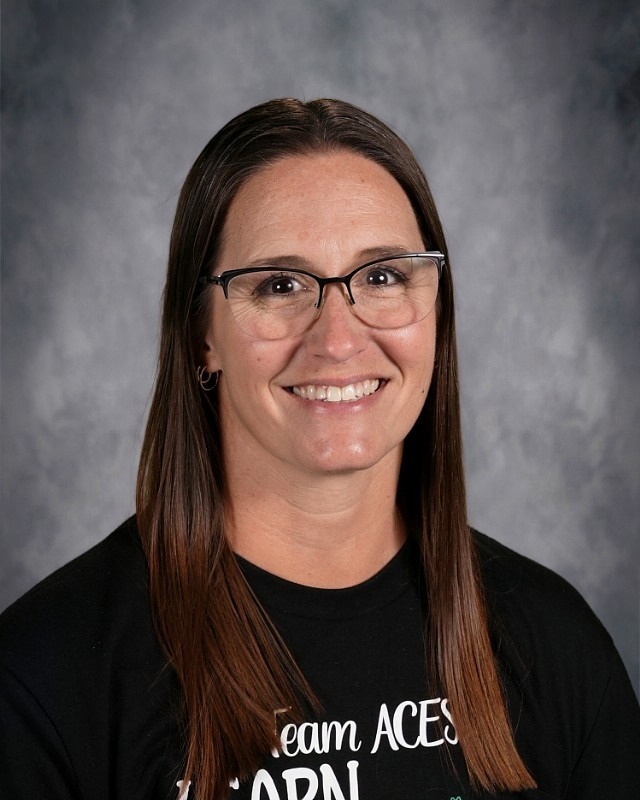 A woman with long brown hair and glasses smiles. She wears a black shirt with "Team ACEs" on it.