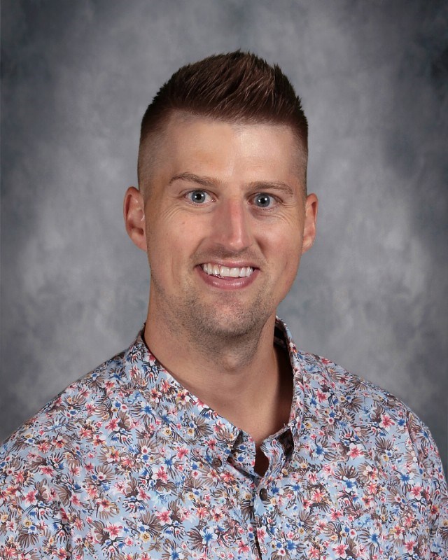 A man wearing a floral shirt smiles brightly at the camera, exuding a cheerful and friendly demeanor.