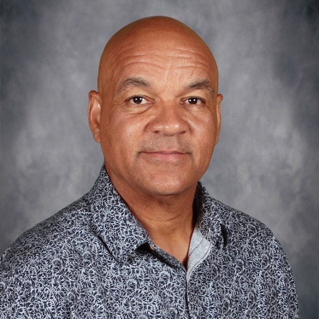 A man wearing a patterned dark shirt poses against a neutral grey background.