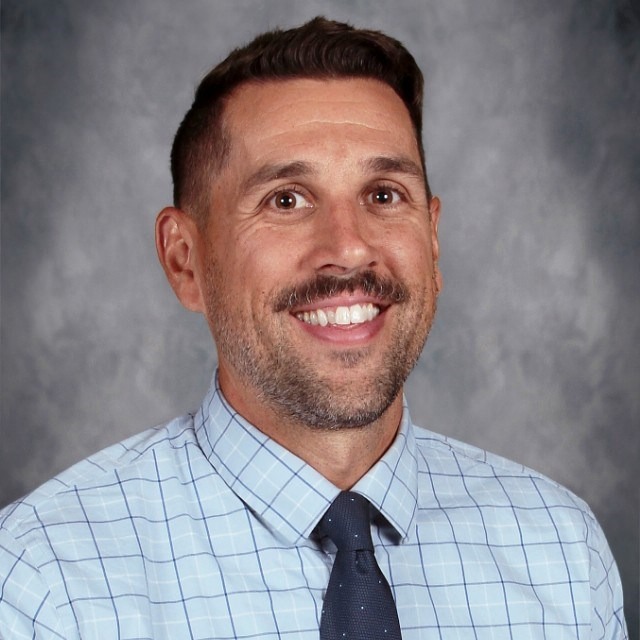 A man in a light blue checked shirt and dark tie poses against a blurred gray backdrop, exuding professionalism and confidence.