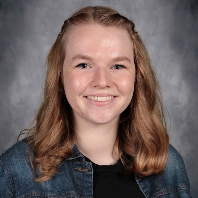 A young woman with long, wavy brown hair wearing a denim jacket and black shirt, posed against a soft gray background.