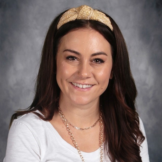 A woman with long, wavy brown hair wears a gold headband and layered necklaces, dressed in a white shirt against a gray background.