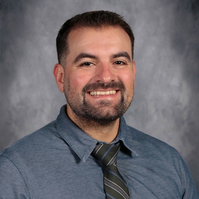 A man wearing a blue button-up shirt and a striped tie, posing against a grey background.