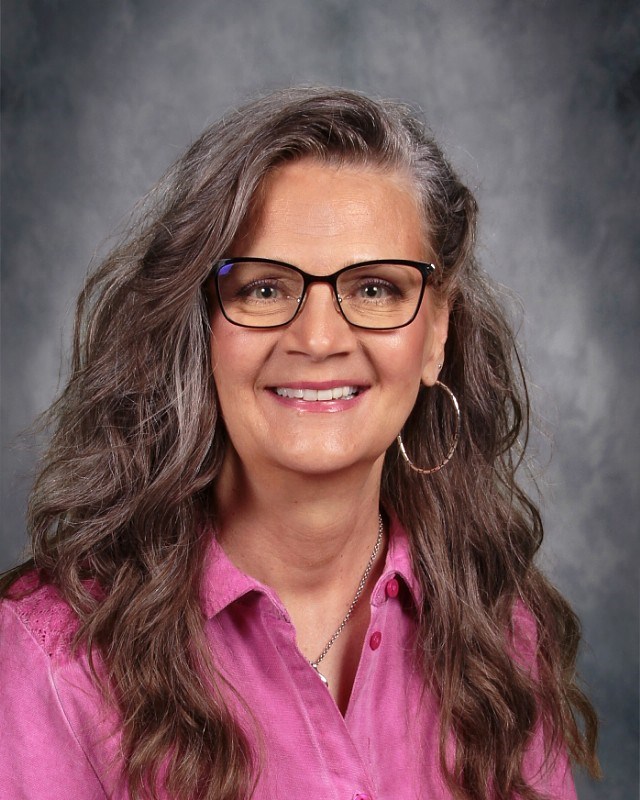 A person with long, wavy dark hair wearing a pink shirt with button details and a silver necklace, set against a gray backdrop.