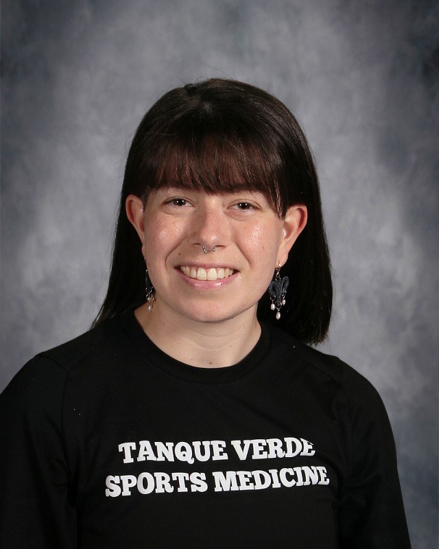A person wearing a black shirt with "Tanque Verde Sports Medicine" printed in white, set against a gray backdrop.