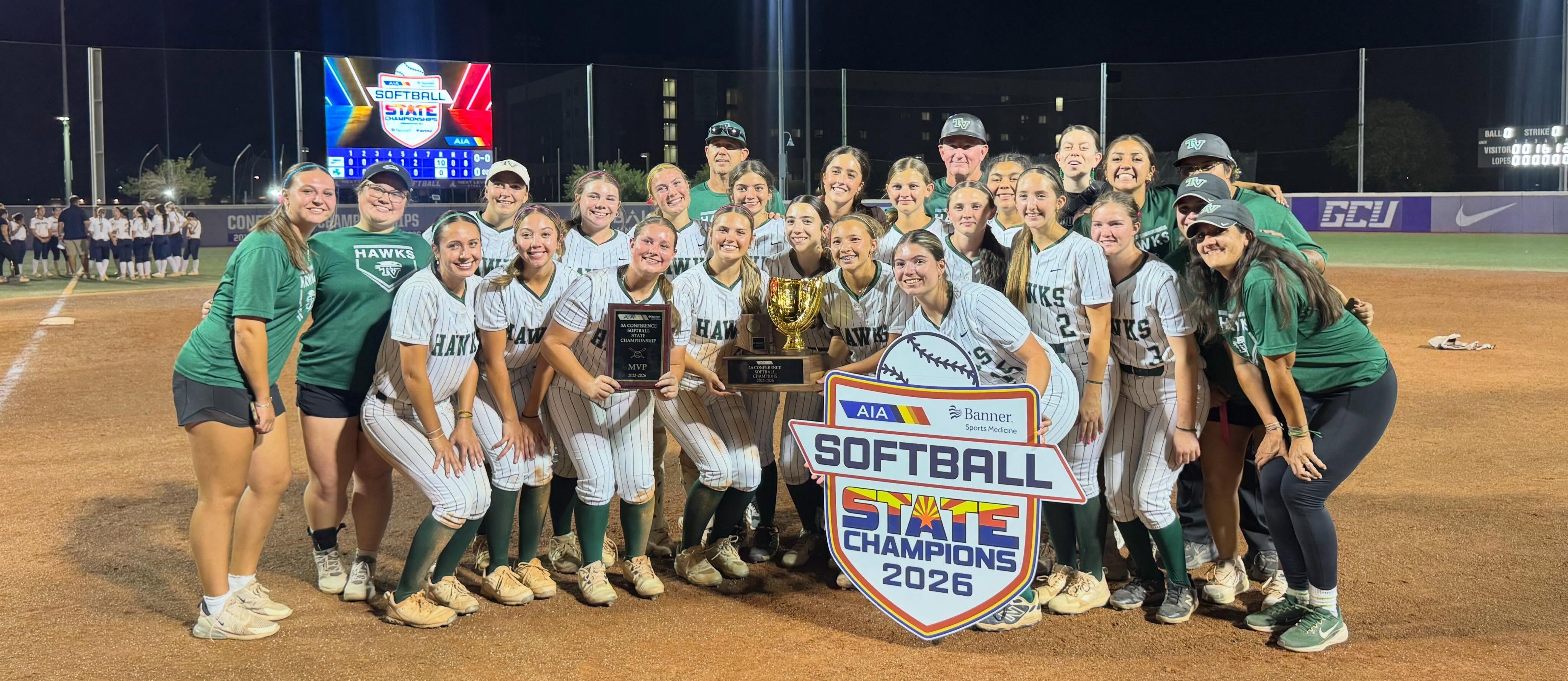 A team of softball players poses for a photo with a trophy on a field at night.