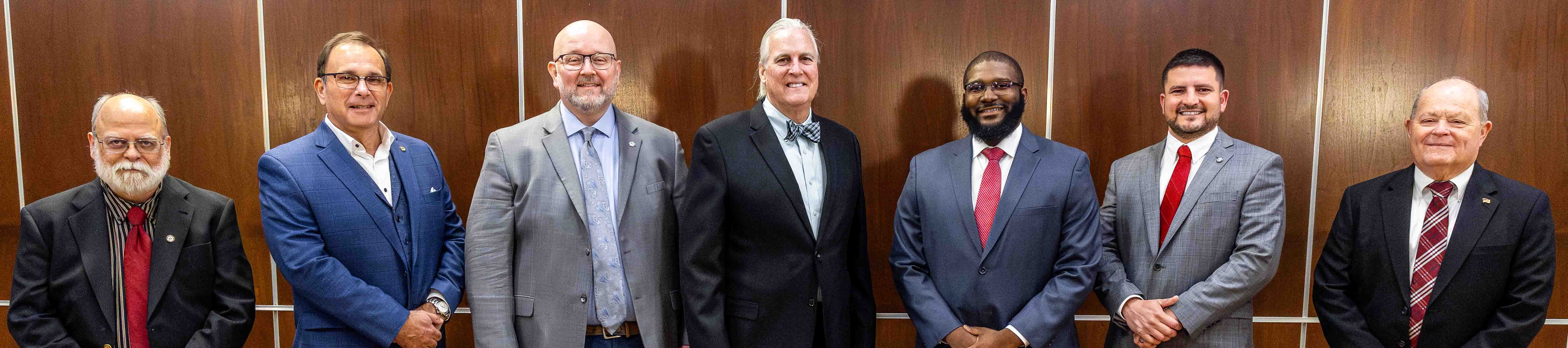 Seven men stand in line dressed in suits and ties in front of a wooden wall backdrop