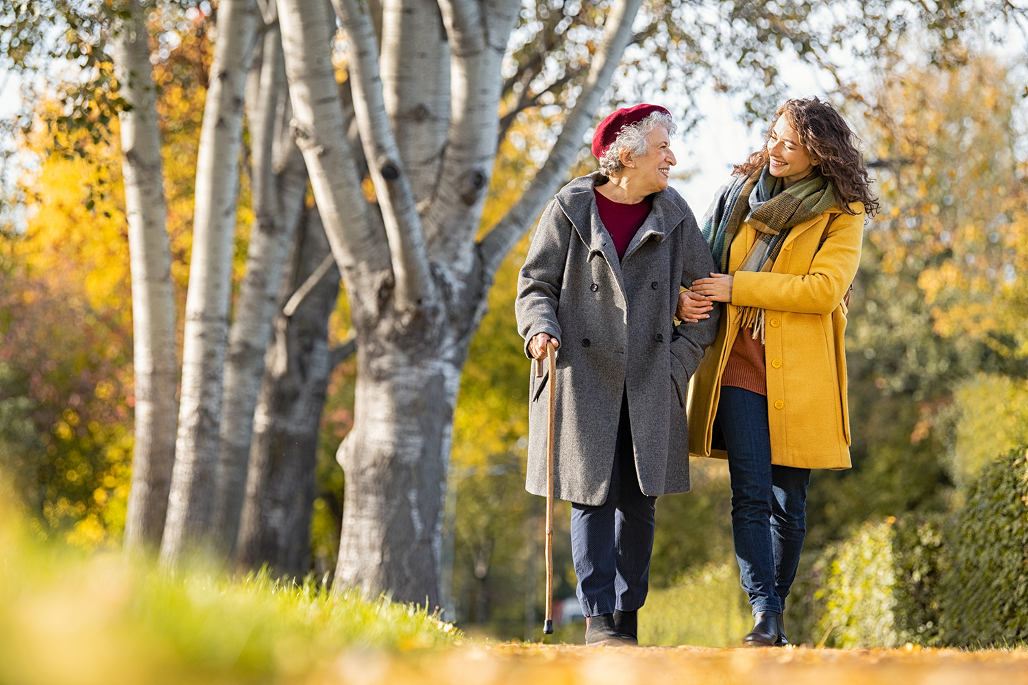 A middle age person helps an elderly person walk. They are wearing autumn attire like hats, scarves and coats.