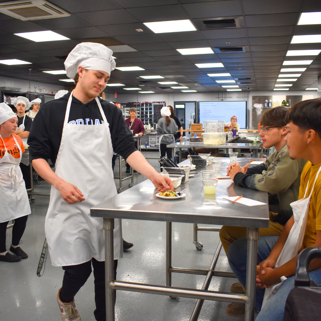 A student handing over a pasta dish to the student judges while others look on