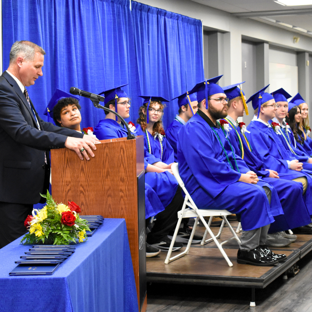 A photo of the graduates on stage from a side angle as the superintendent speaks at the podium
