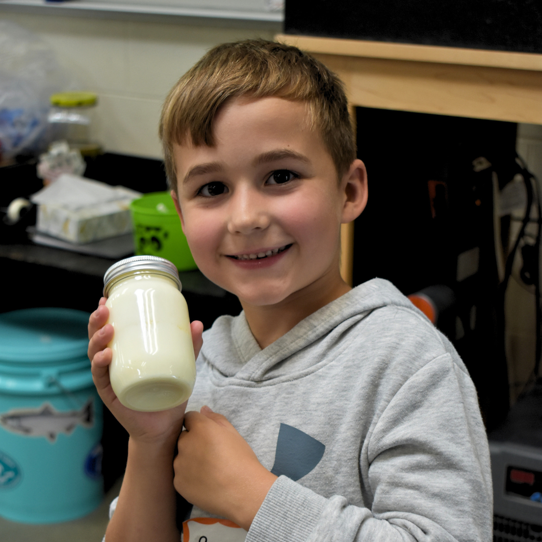 A young student smiling at the camera holding his jar of butter that he made