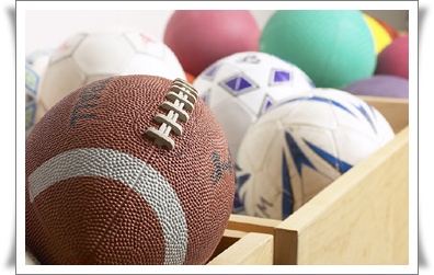 A variety of sports balls, including a football and several colorful balls, are arranged in a wooden box.