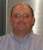 A man with short hair and glasses, wearing a checkered shirt, smiles in front of a blurred background.