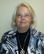 A woman with shoulder-length blonde hair, wearing glasses and a patterned black and white jacket, poses for a portrait against a plain background.