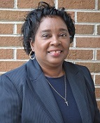 A woman with short, curly hair wearing a dark blazer and a necklace, smiling in front of a brick wall.