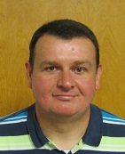 A man with short dark hair and a slight smile, wearing a striped polo shirt, stands in front of a wooden background.