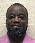 A man with a beard and short hair, wearing a pink shirt, smiles at the camera against a plain background.