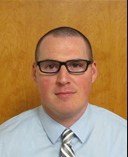 A man with short hair and glasses is wearing a light blue dress shirt and a striped tie, standing in front of a wooden background.