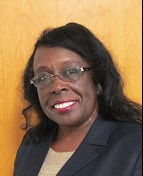 A woman with long black hair and glasses smiles warmly while wearing a dark blazer over a light-colored top, set against a wooden background.