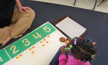 A child with braided hair is engaged in a learning activity on a mat, using colorful counters to count along a number line marked with numbers 1 to 5, while an adult sits nearby.