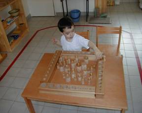 A young child is engaged in play at a wooden table, manipulating various wooden blocks and pieces arranged in a structured layout.