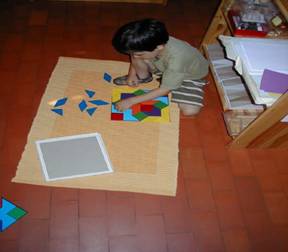 A child is sitting on a mat, arranging colorful geometric shapes on the floor, with a blank square and other materials nearby.