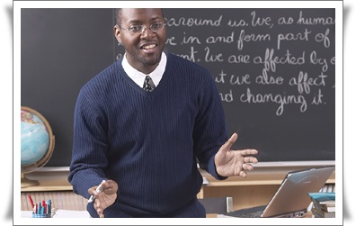 A smiling teacher stands in front of a chalkboard, gesturing as he explains a lesson, with a globe and desk visible in the classroom.