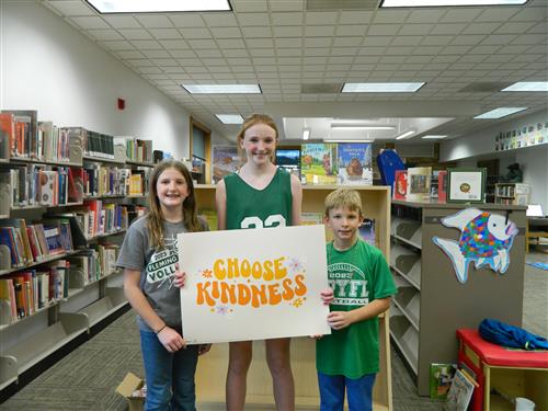 Three children in a library with a sign that reads "choose kindness.