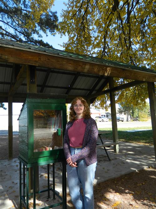 A young woman stands next to a small, outdoor wooden structure, perhaps part of a park or public space.