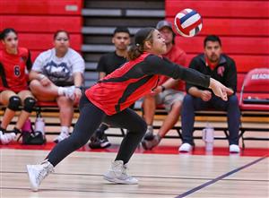 A girl in a red and black sports uniform is playing volleyball, reaching up to hit the ball.