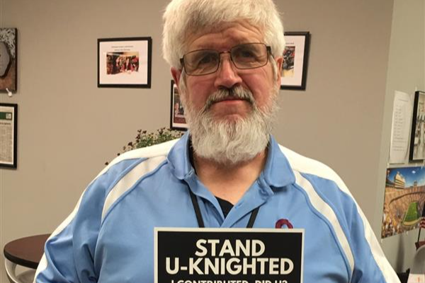 An individual standing in an office, proudly holding a sign that reads 'Stand U-Knighted. I contributed. Did you?'