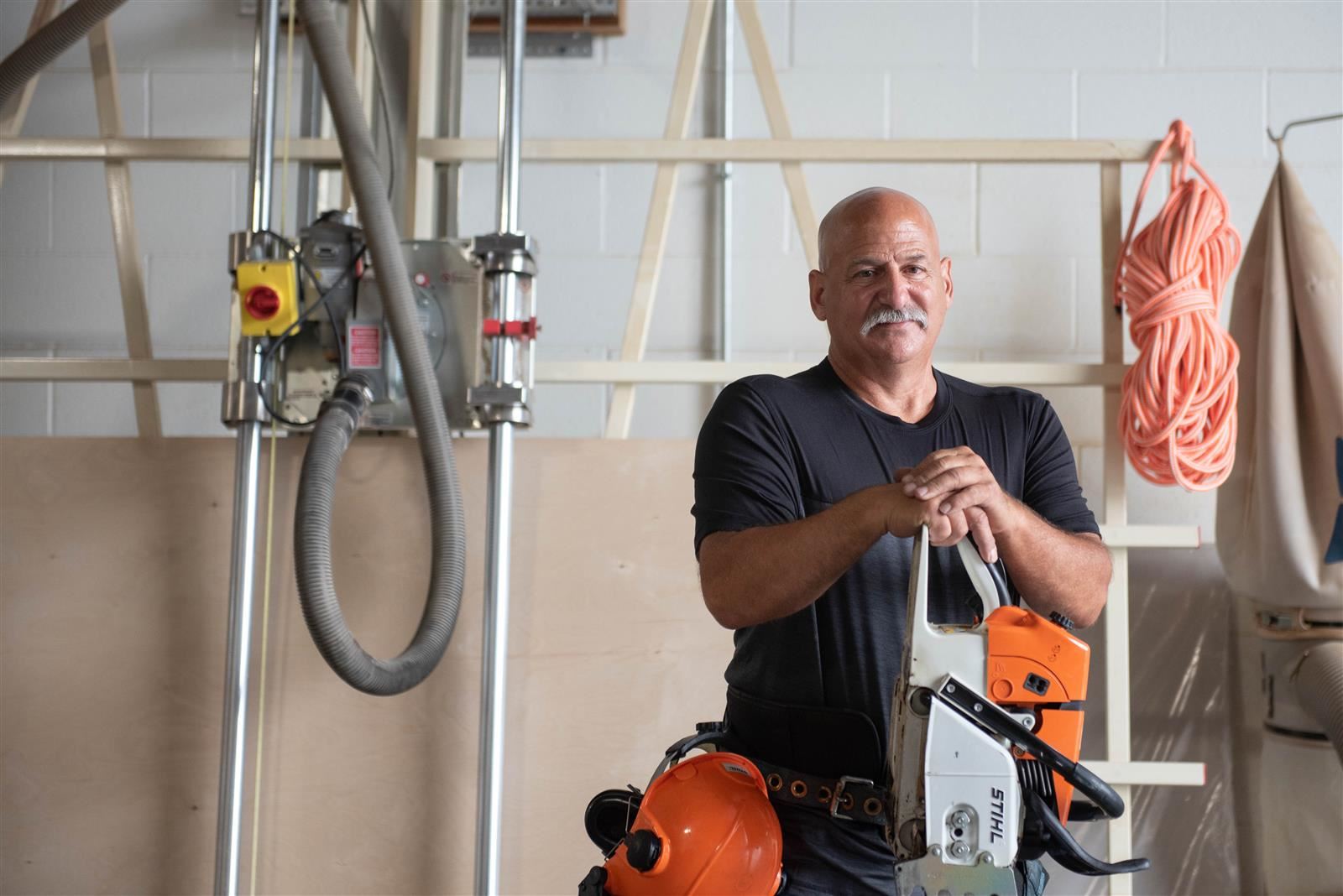 A man in a workshop, leaning against the workbench, surrounded by tools and equipment.