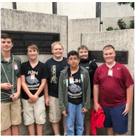 A group of students posing for a photo in front of a building, standing in front of a plaque or sign.
