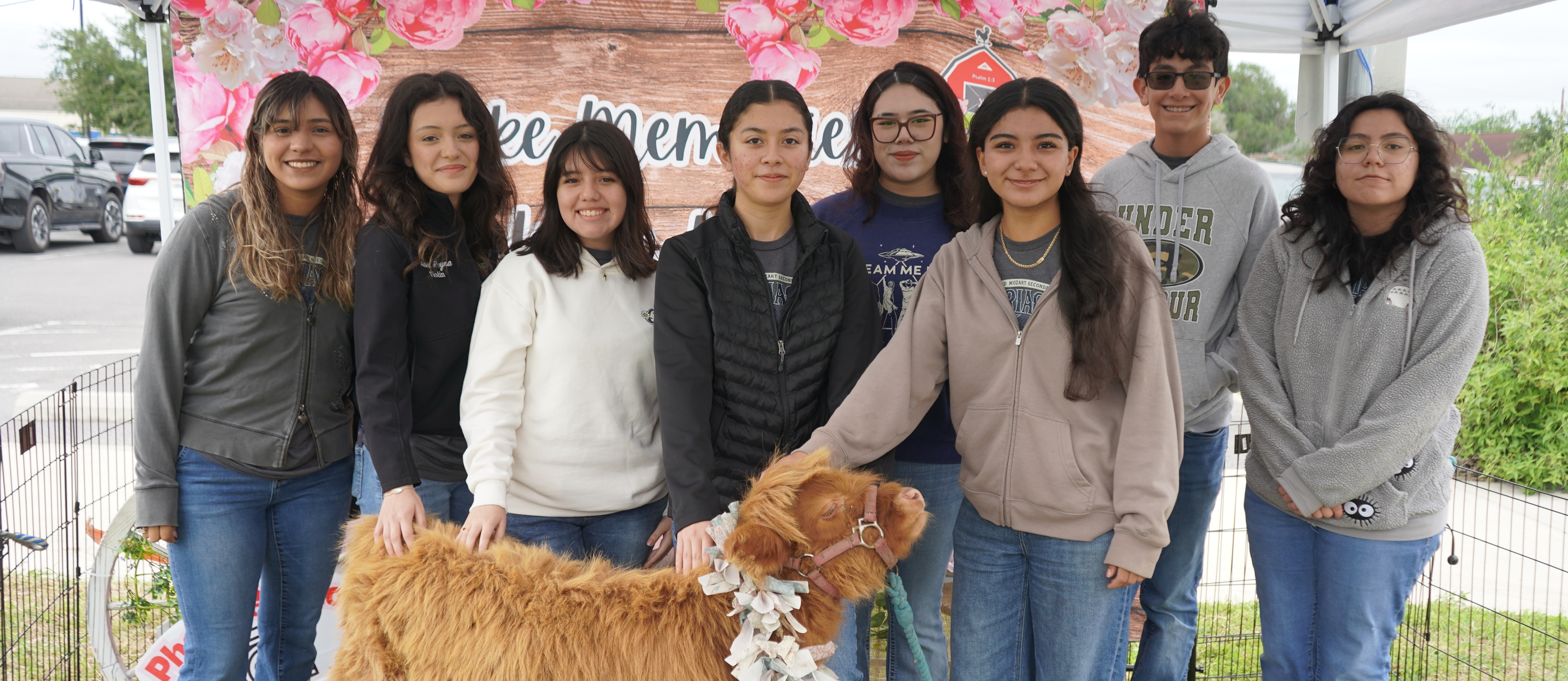 A group of people stands under a tent, posing with a light brown sheep. Some wear jackets, jeans, and sneakers.