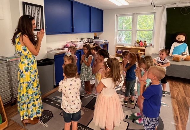 A teacher in a floral dress is leading a group of children in a classroom setting, with various educational materials and a playful atmosphere.