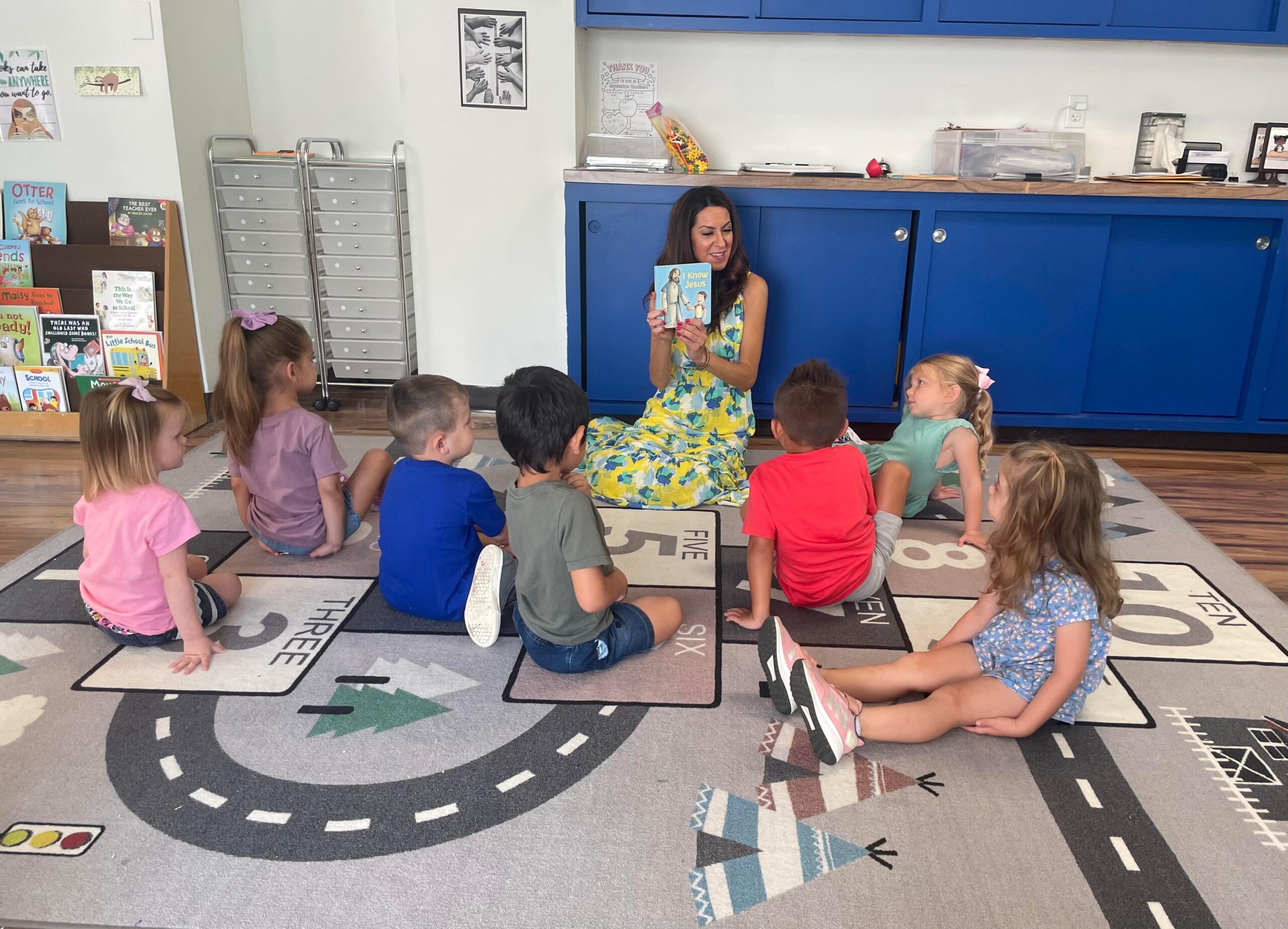 A teacher reads a book to a group of children sitting on a colorful rug in a classroom setting.