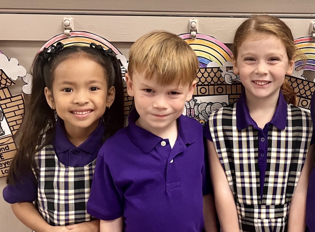 Three children, two girls and one boy, are smiling together in front of a colorful backdrop featuring rainbows and clouds, all wearing matching purple and plaid outfits.