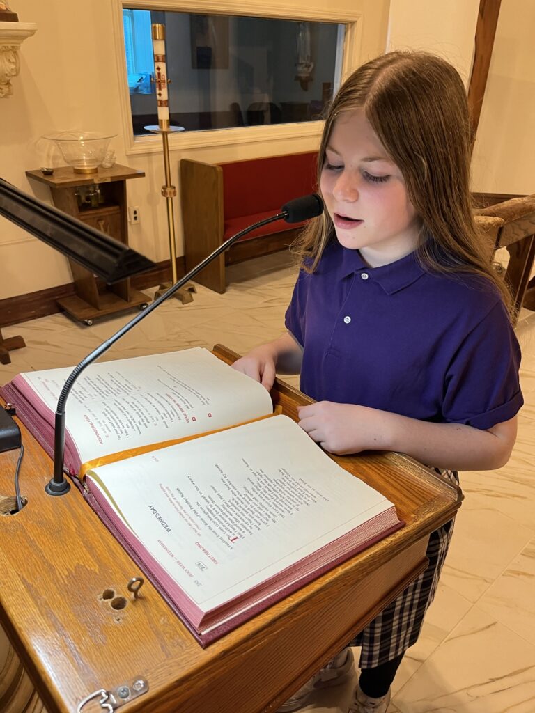 A young girl in a purple shirt stands at a lectern, reading from an open book with a microphone in front of her.