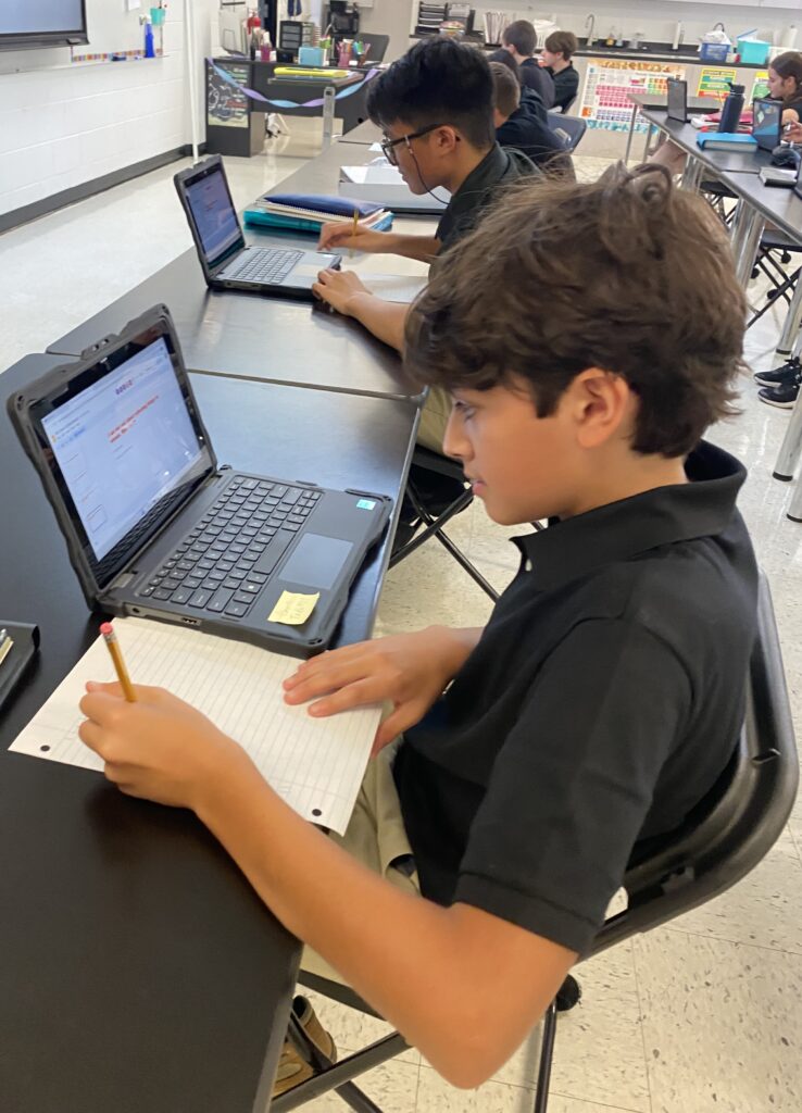 A student in a black shirt is focused on writing on a sheet of paper while using a laptop in a classroom setting, with another student visible in the background.