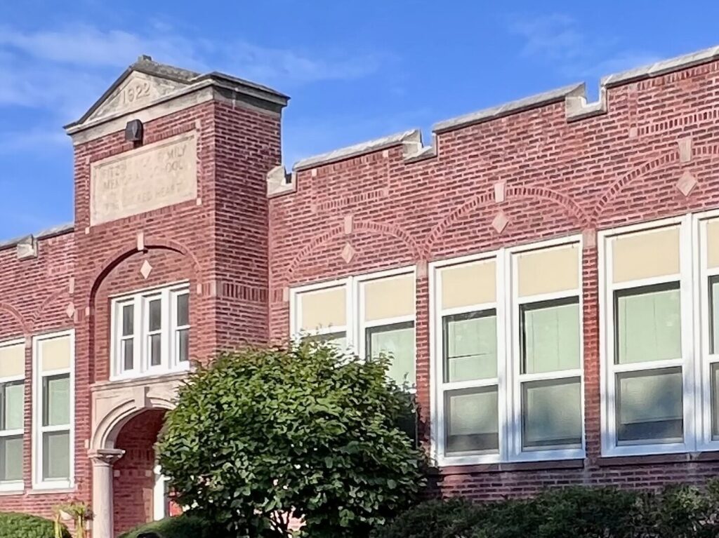 A brick building with large windows and a decorative entrance, surrounded by greenery under a clear blue sky.
