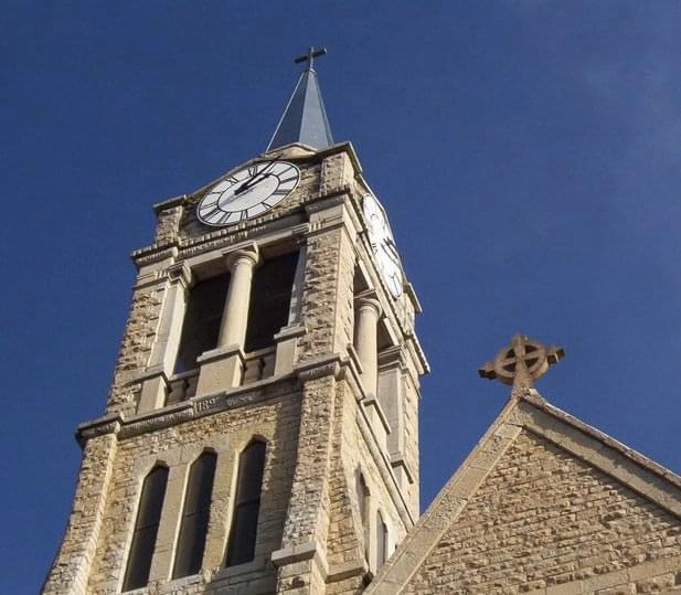 A close-up view of a church tower featuring a clock and a cross, set against a clear blue sky.