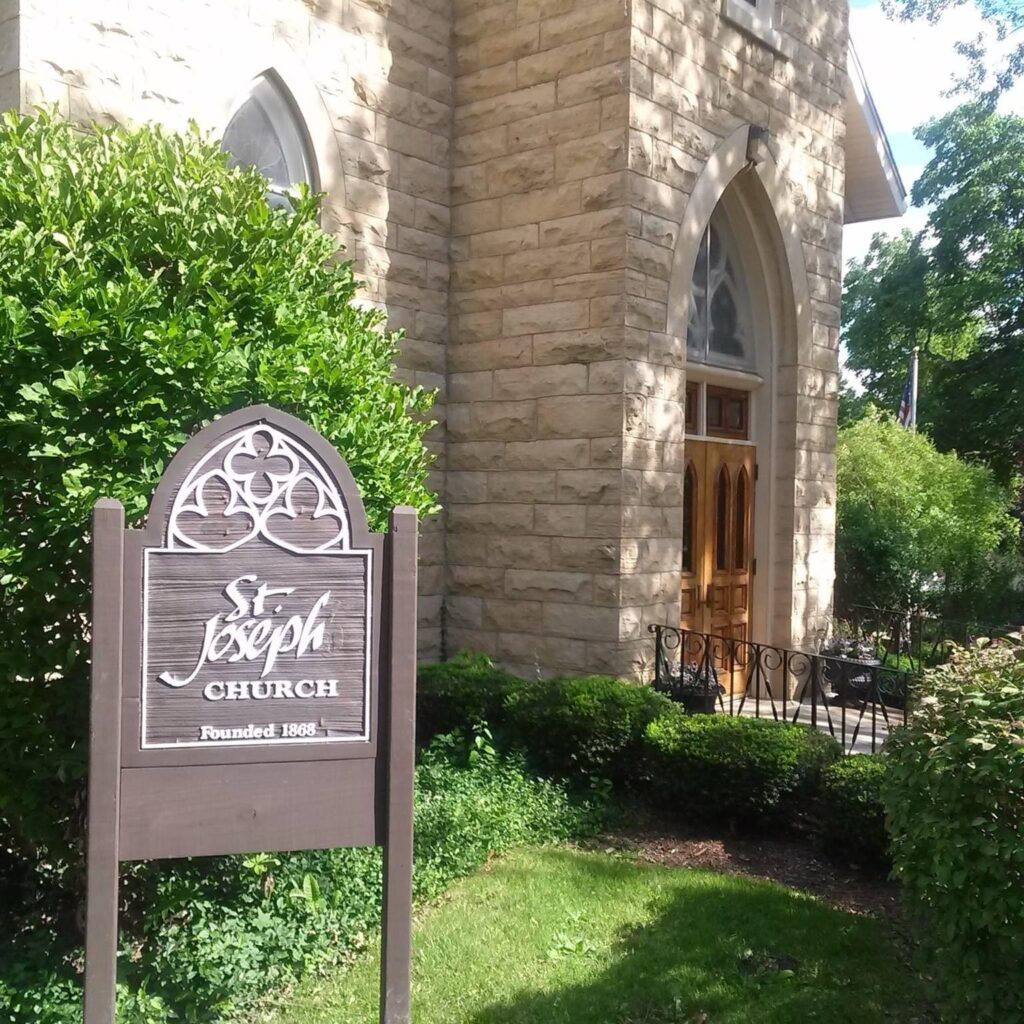 A sign for St. Joseph Church stands in front of a stone church building, surrounded by greenery.