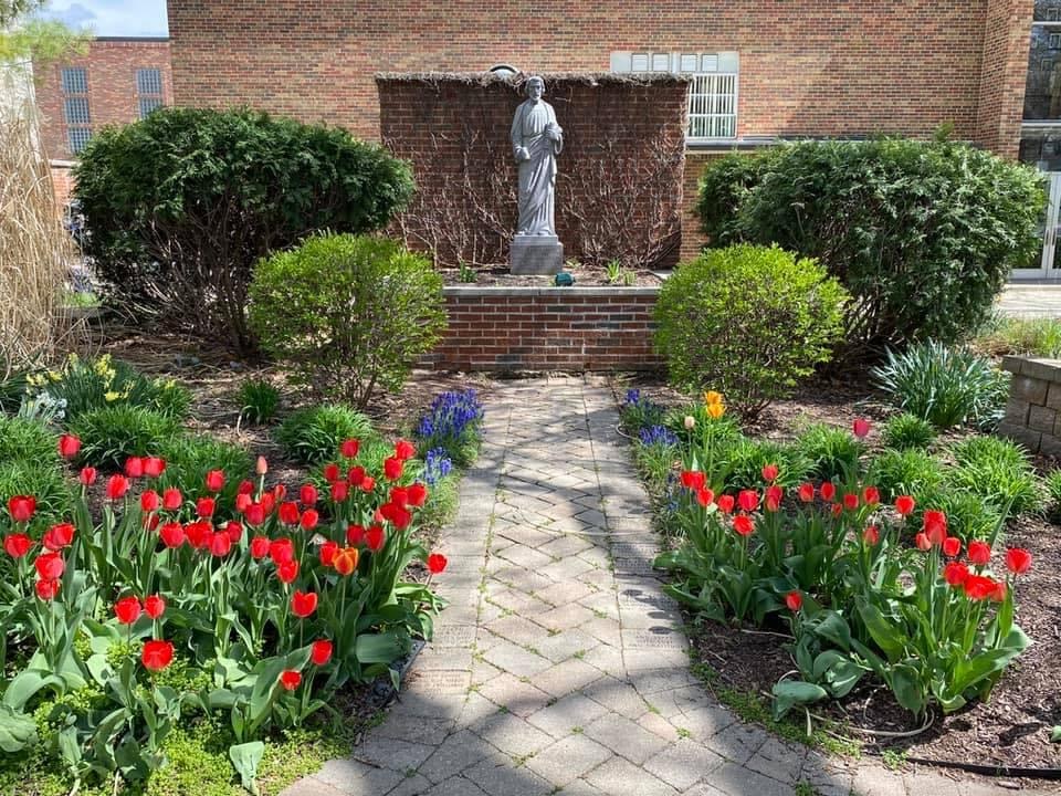 A landscaped garden featuring vibrant red tulips, green shrubs, and a stone statue at the center, with a brick wall in the background.