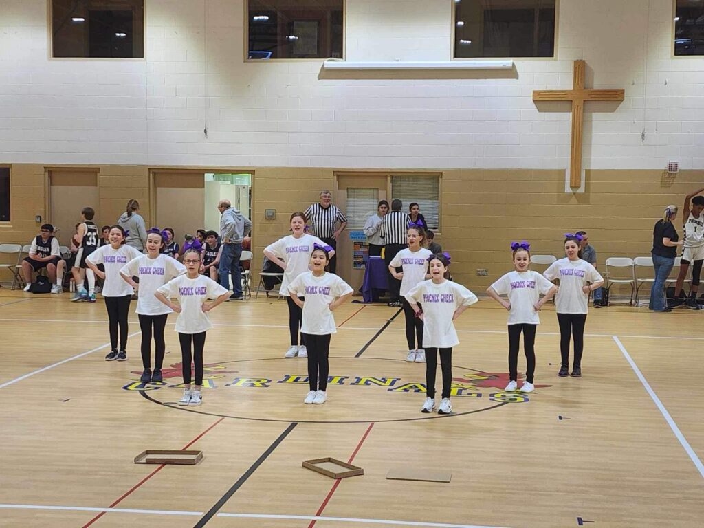 A group of young cheerleaders in matching white shirts and black leggings perform in a gymnasium, with a wooden cross visible in the background.