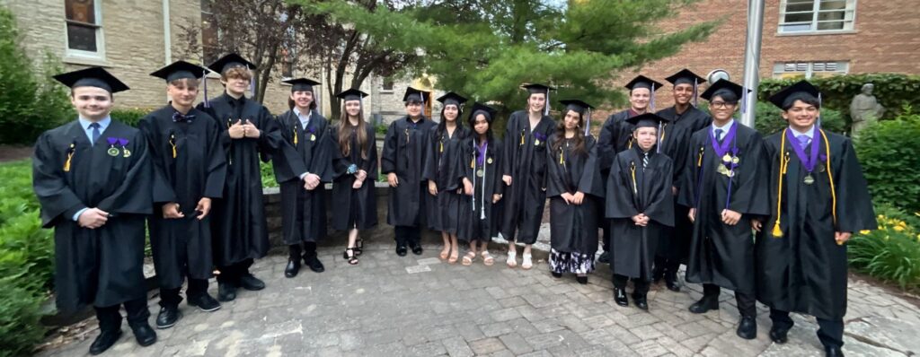 A group of graduates in black caps and gowns stands together outdoors, smiling and celebrating their achievement.