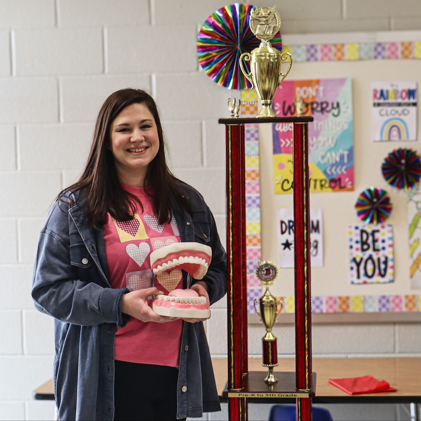 Lauren Mitchell, speech-language pathologist serving Gleason and Greenfield Schools, smiles while standing next to the tall traveling Feature Teacher trophy in a classroom. She is holding a model of teeth used for speech instruction, with colorful classroom posters visible behind her.