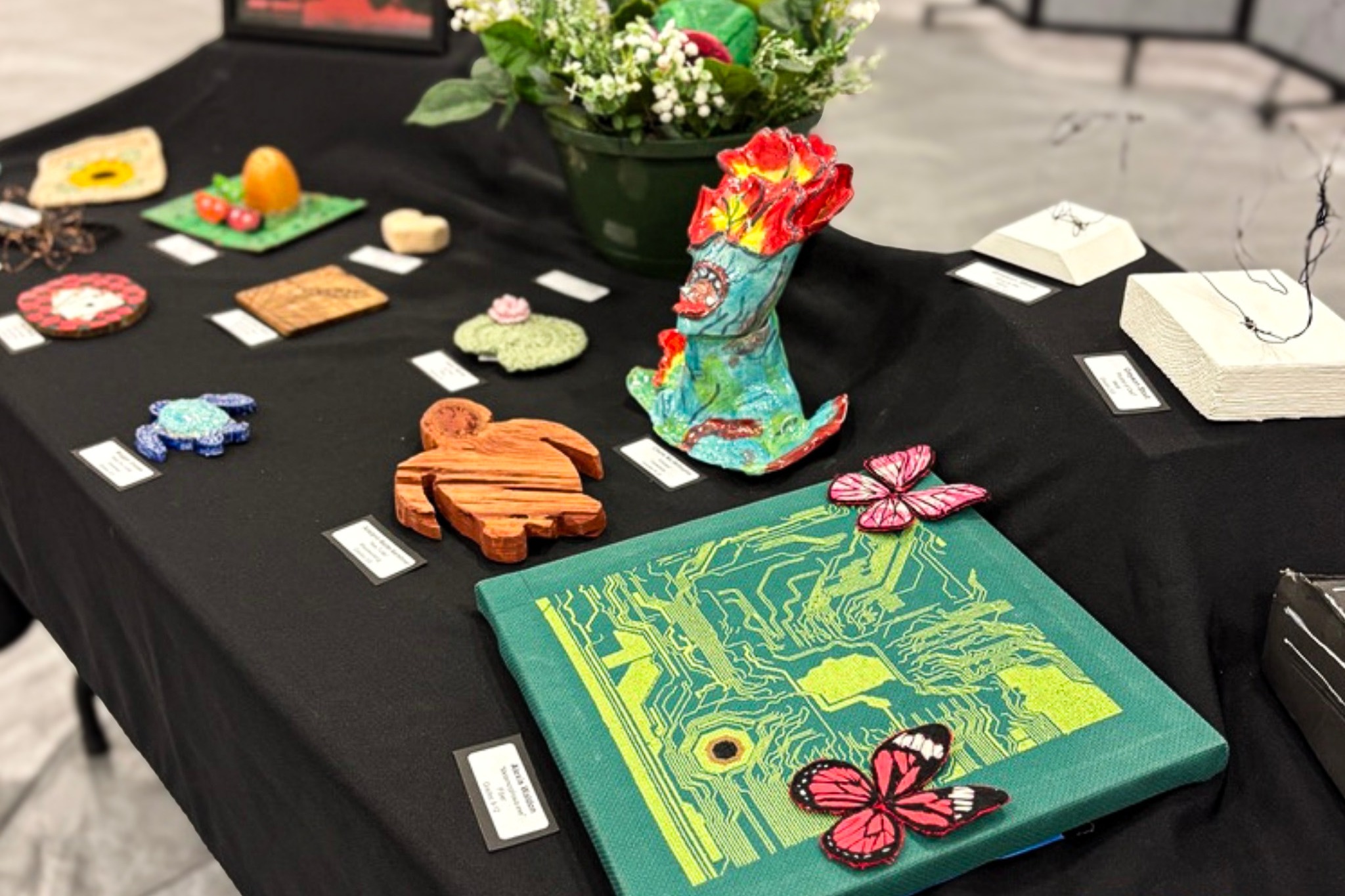 A table with a black tablecloth displays various art pieces, including sculptures, a canvas with a butterfly design, and a flower vase.
