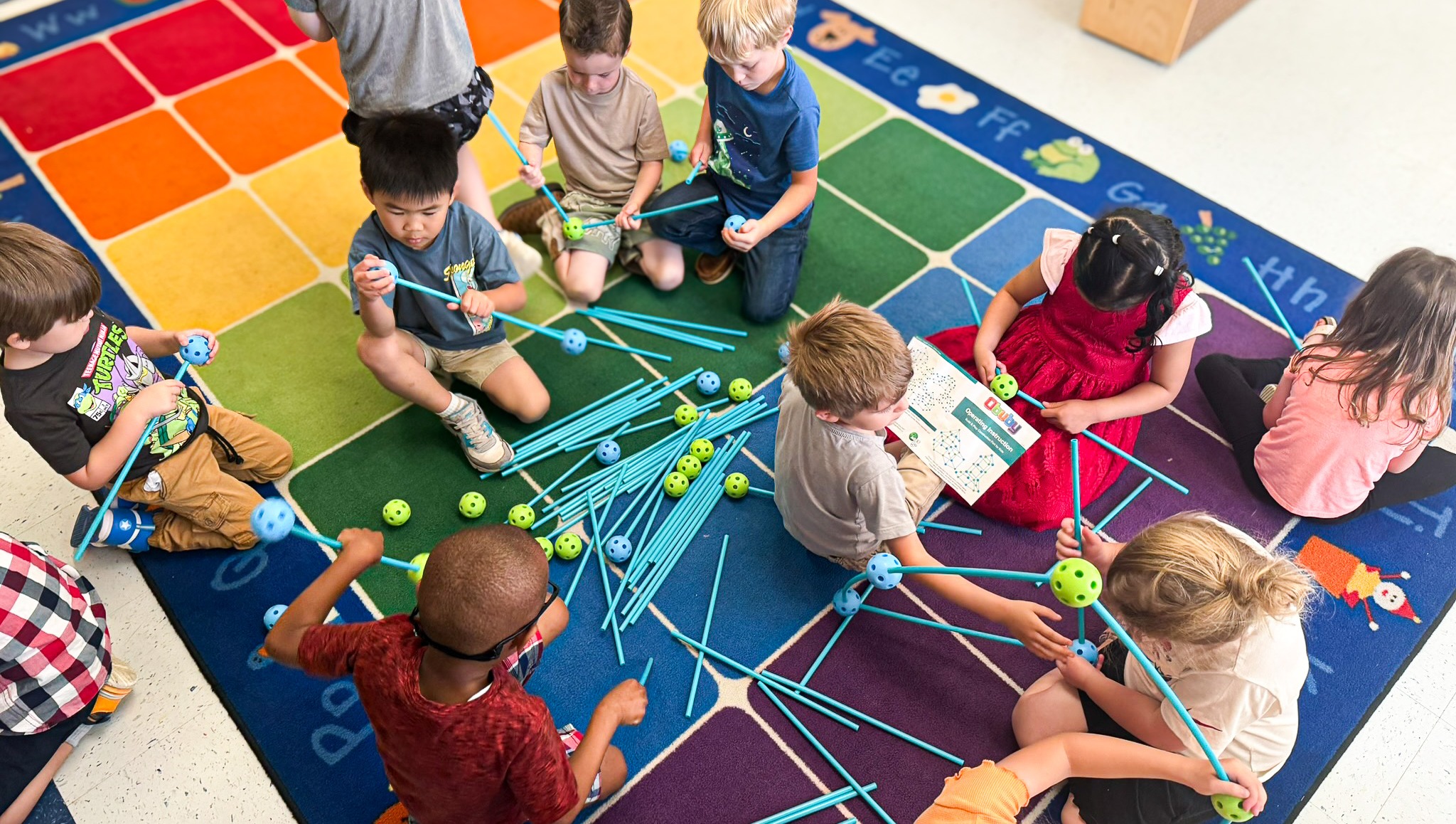 Group of children sitting on floor, holding rods, and assembling them into a structure.