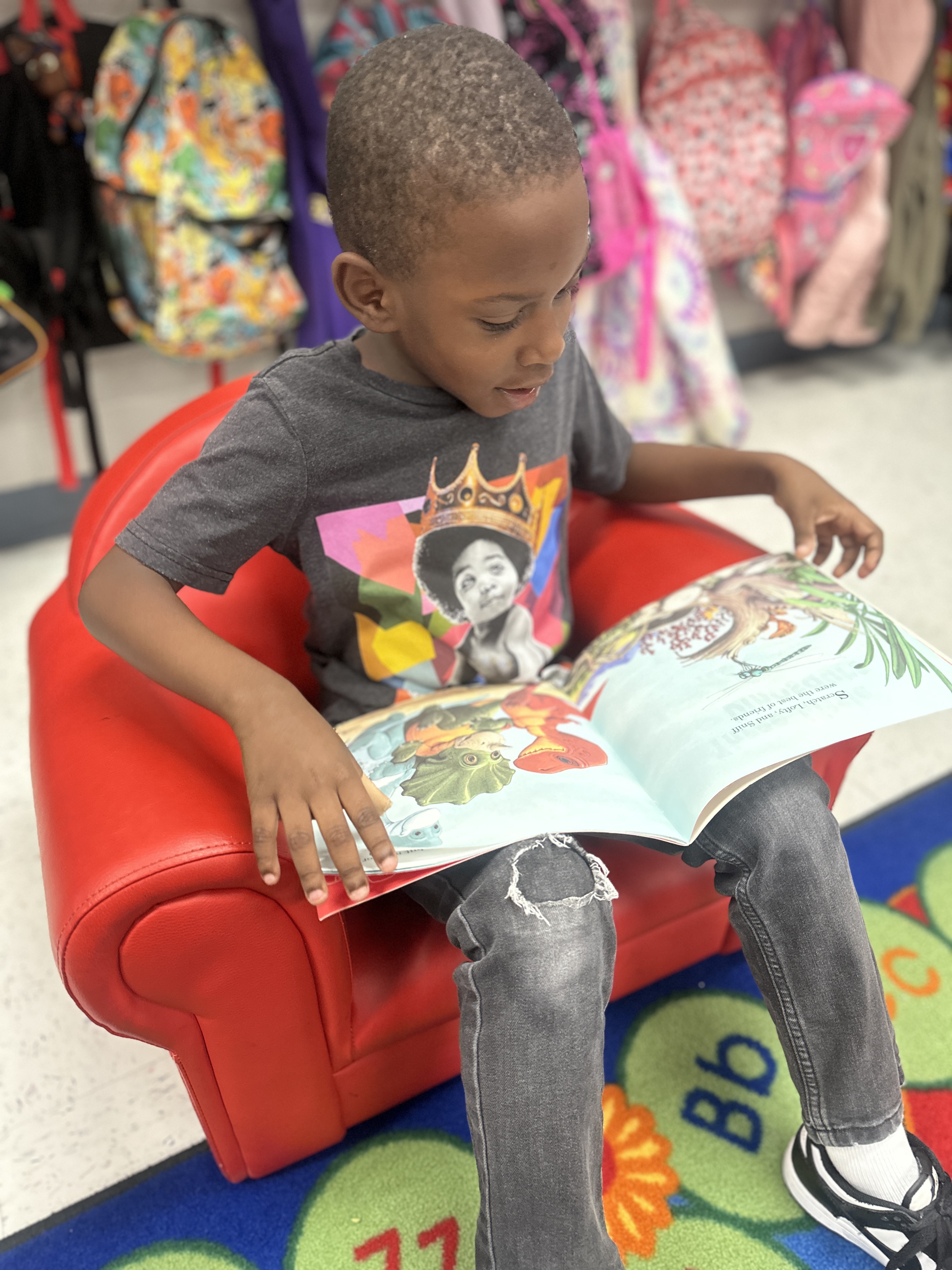A child in a chair, reading a book with illustrations, surrounded by colorful backpacks.
