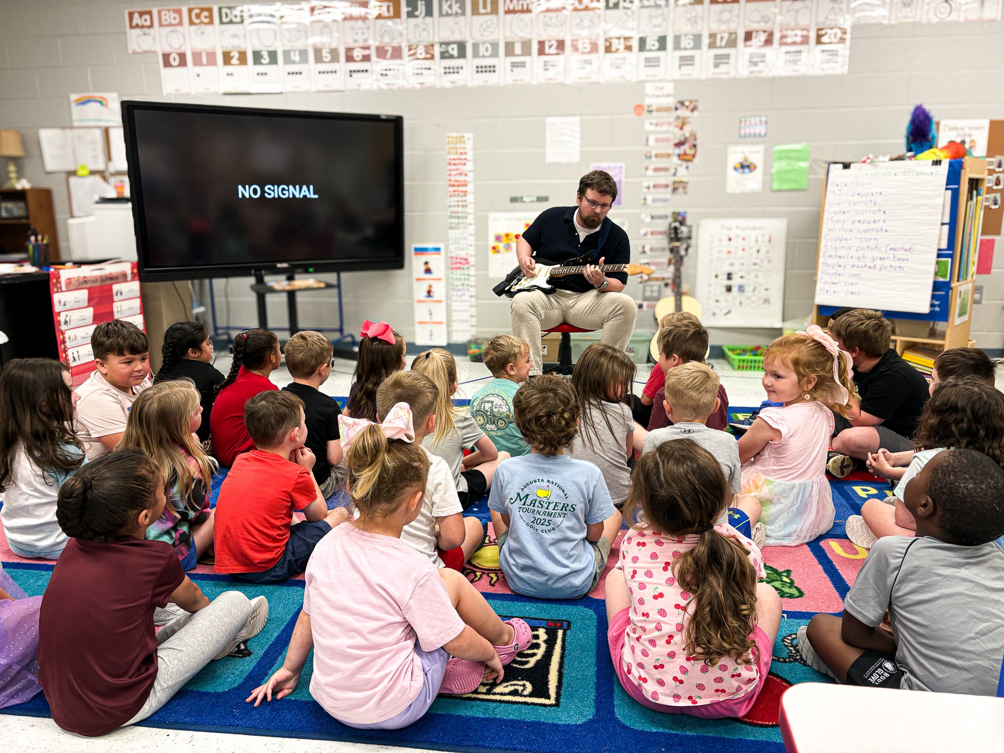 A man plays a guitar in a classroom; children sit in a circle around him, watching.